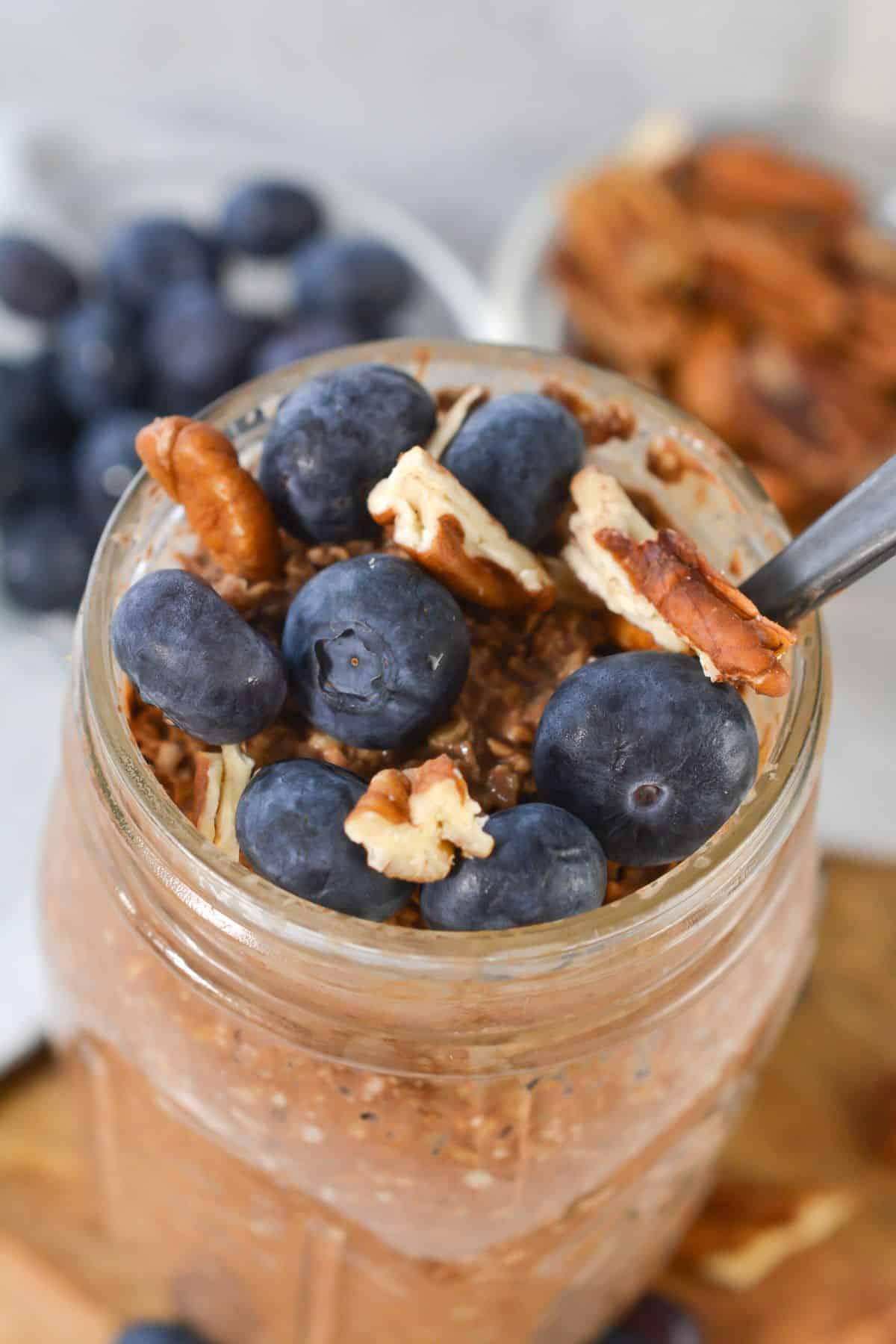 A mason jar filled with tiramisu overnight oats, topped with fresh blueberries and pecan pieces, with a spoon inside.