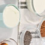 Assorted ingredients in glass bowls and cups on a white surface, including yogurt, milk, coffee, chia seeds, cocoa powder, and a brown liquid—perfect for making Tiramisu Overnight Oats.