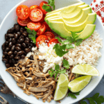 A bowl filled with meat, black beans, cherry tomatoes, avocado slices, rice, lime wedges, crumbled cheese, and cilantro leaves. Forks are placed beside the bowl.