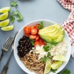 A bowl featuring shredded meat, black beans, cherry tomatoes, avocado slices, rice, lime wedges, and cheese crumbles in a bowl, with tortillas, lime slices, and utensils on the side.