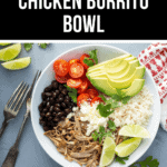A white bowl containing a savory chicken burrito bowl with black beans, shredded chicken, cherry tomatoes, avocado slices, lime wedges, rice, and fresh cilantro. Two forks lie beside the bowl.