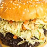 A close-up of an Asian Burger with a sesame seed bun, topped with creamy coleslaw and a thick beef patty.