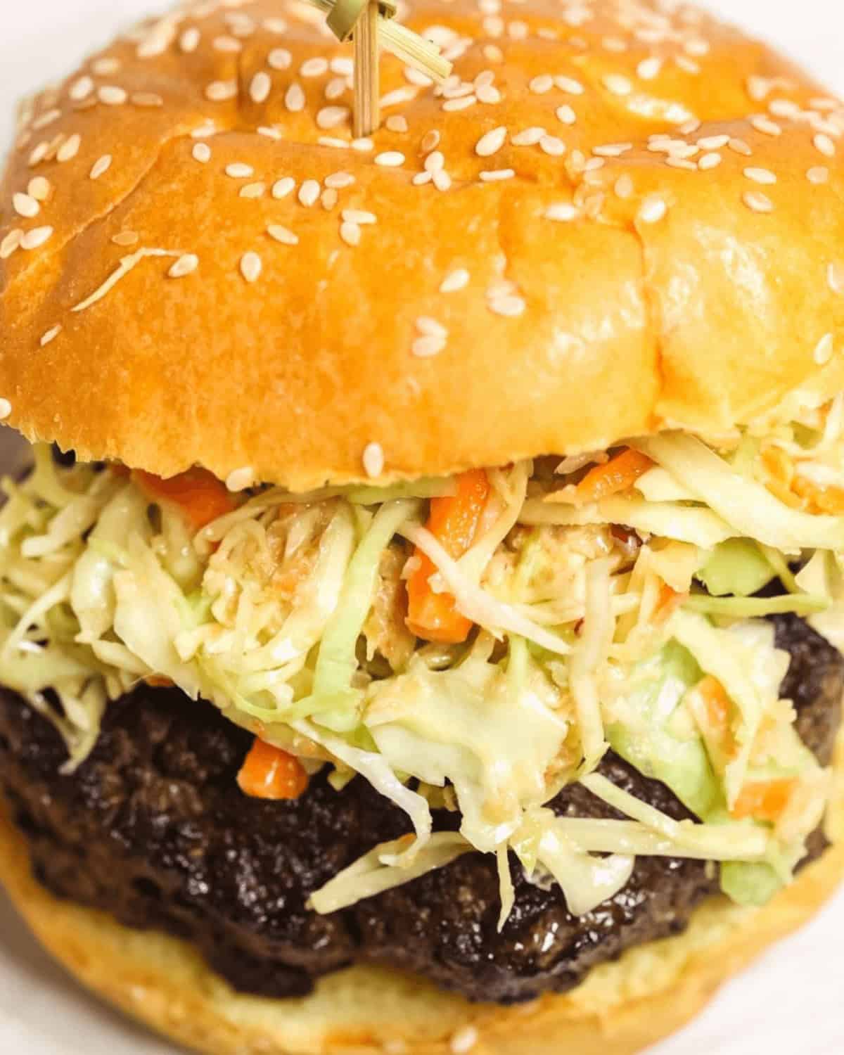 A close-up of an Asian Burger with a sesame seed bun, topped with creamy coleslaw and a thick beef patty.