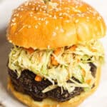 A close-up of an Asian Burger featuring a sesame seed bun, a beef patty, and a generous serving of coleslaw, secured with a wooden skewer.