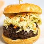 A close-up of an Asian Burger topped with shredded cabbage and carrot slaw, served in a sesame seed bun with a wooden skewer.
