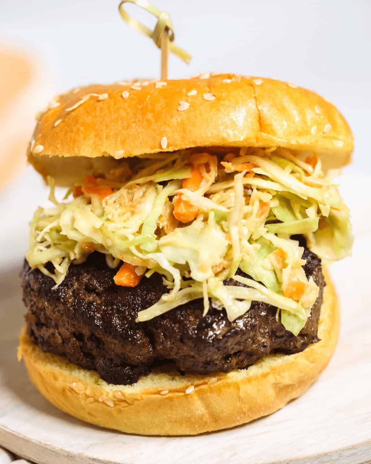 A close-up of an Asian Burger topped with shredded cabbage and carrot slaw, served in a sesame seed bun with a wooden skewer.