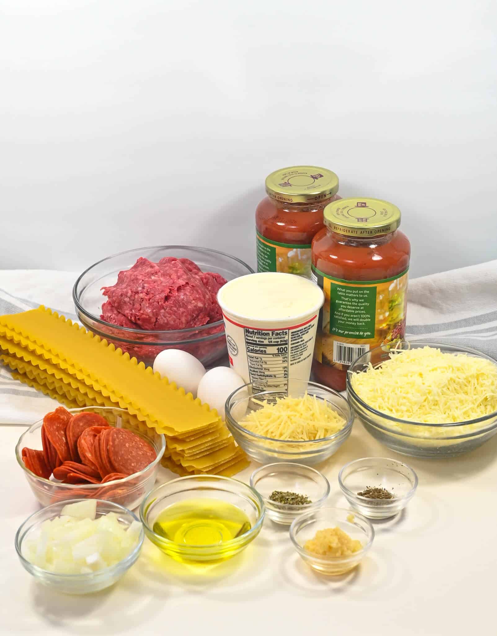Various ingredients for making lazy lasagna arranged on a table, including lasagna noodles, minced meat, tomato sauce, cheeses, eggs, sliced sausage, and spices.