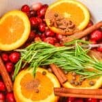 Sliced oranges, fresh cranberries, rosemary sprigs, cinnamon sticks, and whole spices are arranged in a white pot for a festive Christmas Simmering Pot.