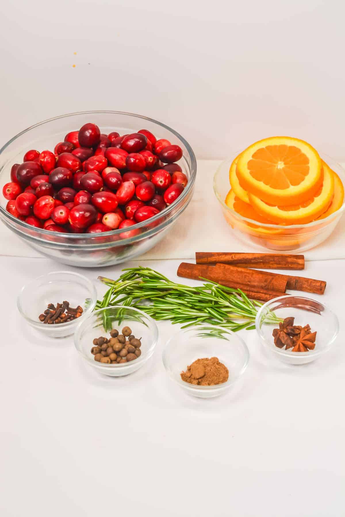 A glass bowl of fresh cranberries, a bowl of orange slices, cinnamon sticks, rosemary sprigs, and small bowls of various spices are arranged on a white surface.