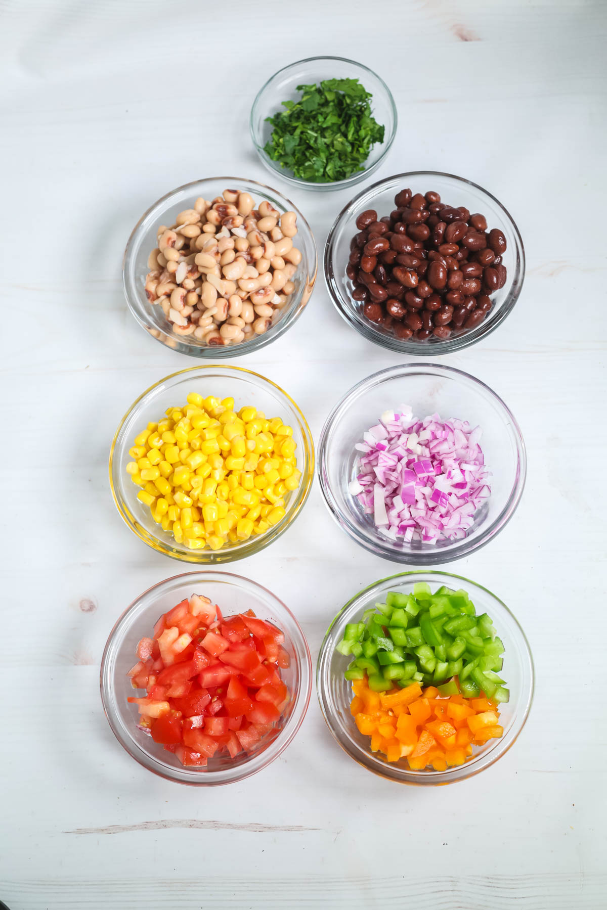Top-down view of various ingredients in bowls of black and white beans, corn, chopped red onions, tomatoes, and bell peppers, on a white surface.