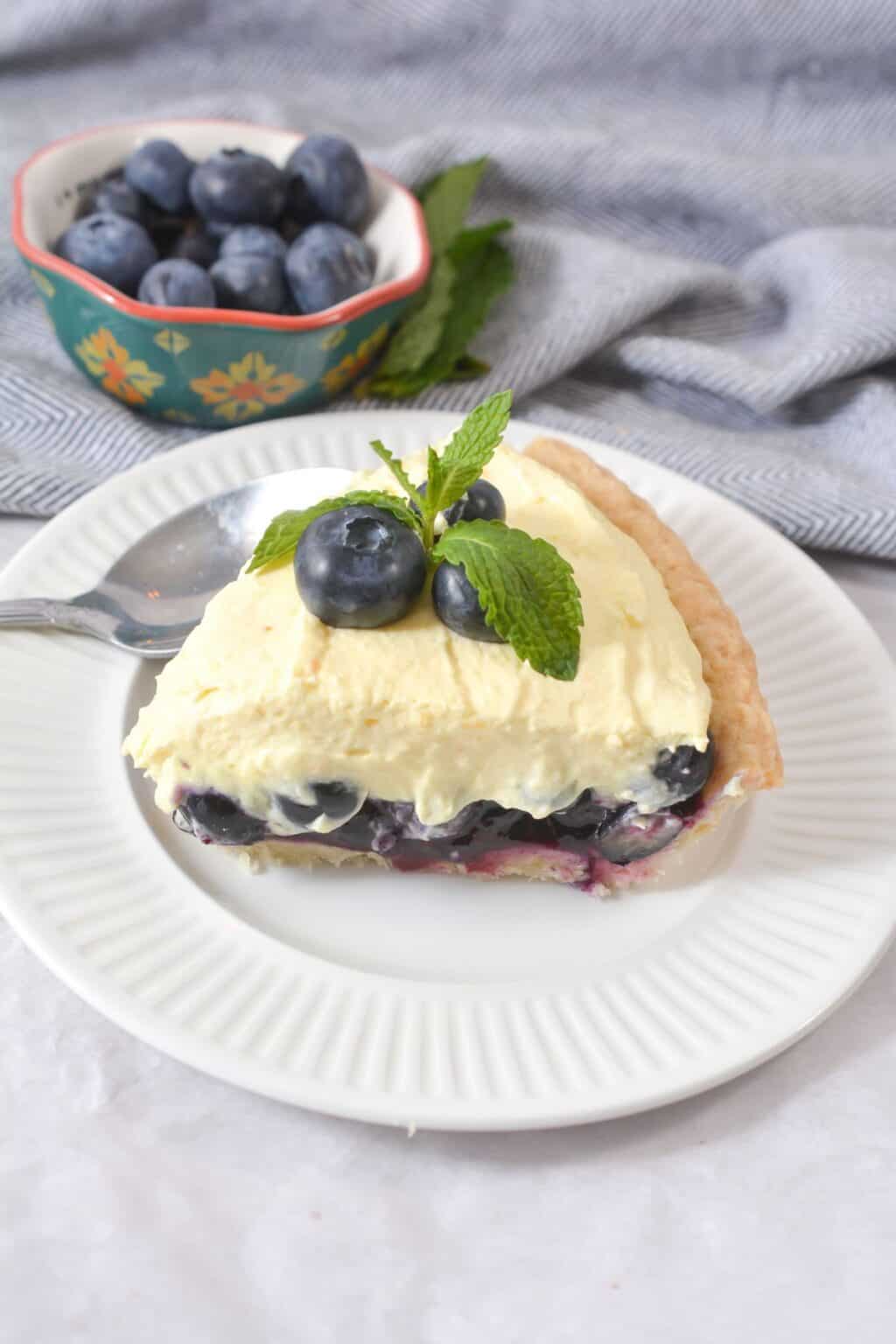 A slice of Blueberry Cream Pie topped with fresh blueberries and mint, served on a white plate with a spoon. In the background, a bowl of plump blueberries completes this tempting dessert scene.