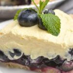 A slice of Blueberry Cream Pie topped with fresh blueberries and a sprig of mint sits elegantly on a white plate.