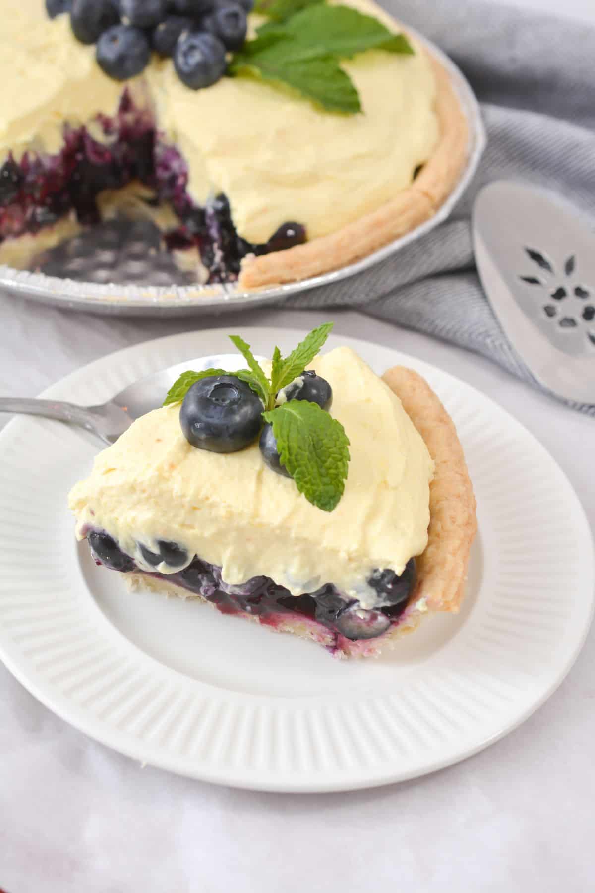 A slice  topped with whipped cream, fresh fruit, and mint leaves sits on a white plate.