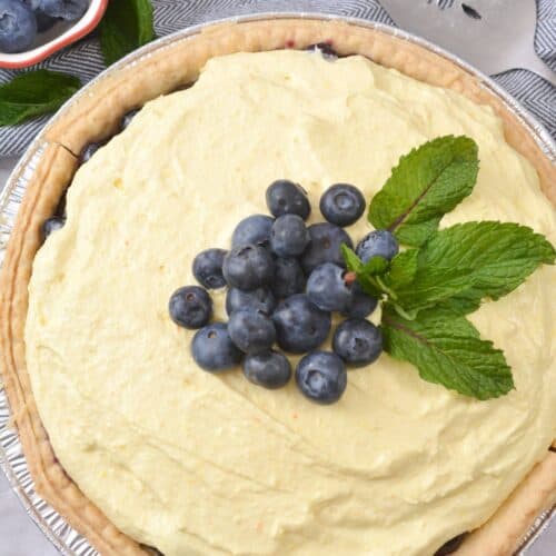 A sumptuous Blueberry Cream Pie topped with fresh blueberries and mint leaves, arranged on a light cloth beside a serving utensil and a small bowl of blueberries.