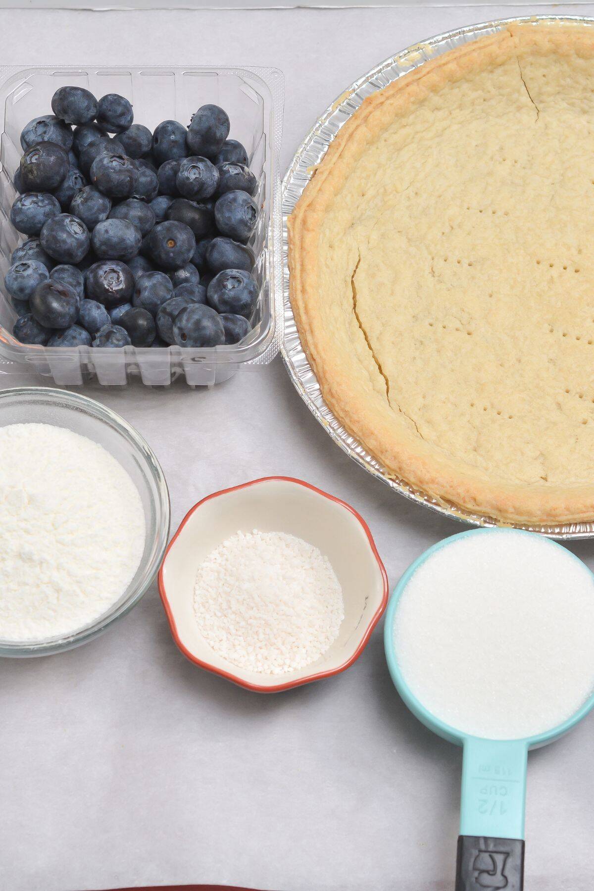 A baked pie crust, fresh blueberries, shredded coconut, granulated sugar, and a small bowl of flour arranged on a light surface—perfect beginnings for making a delicious Blueberry Cream Pie.