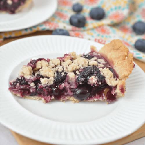 A close-up of a slice of Blueberry Crumb Pie on a white plate. The background includes a patterned cloth and scattered fresh blueberries.