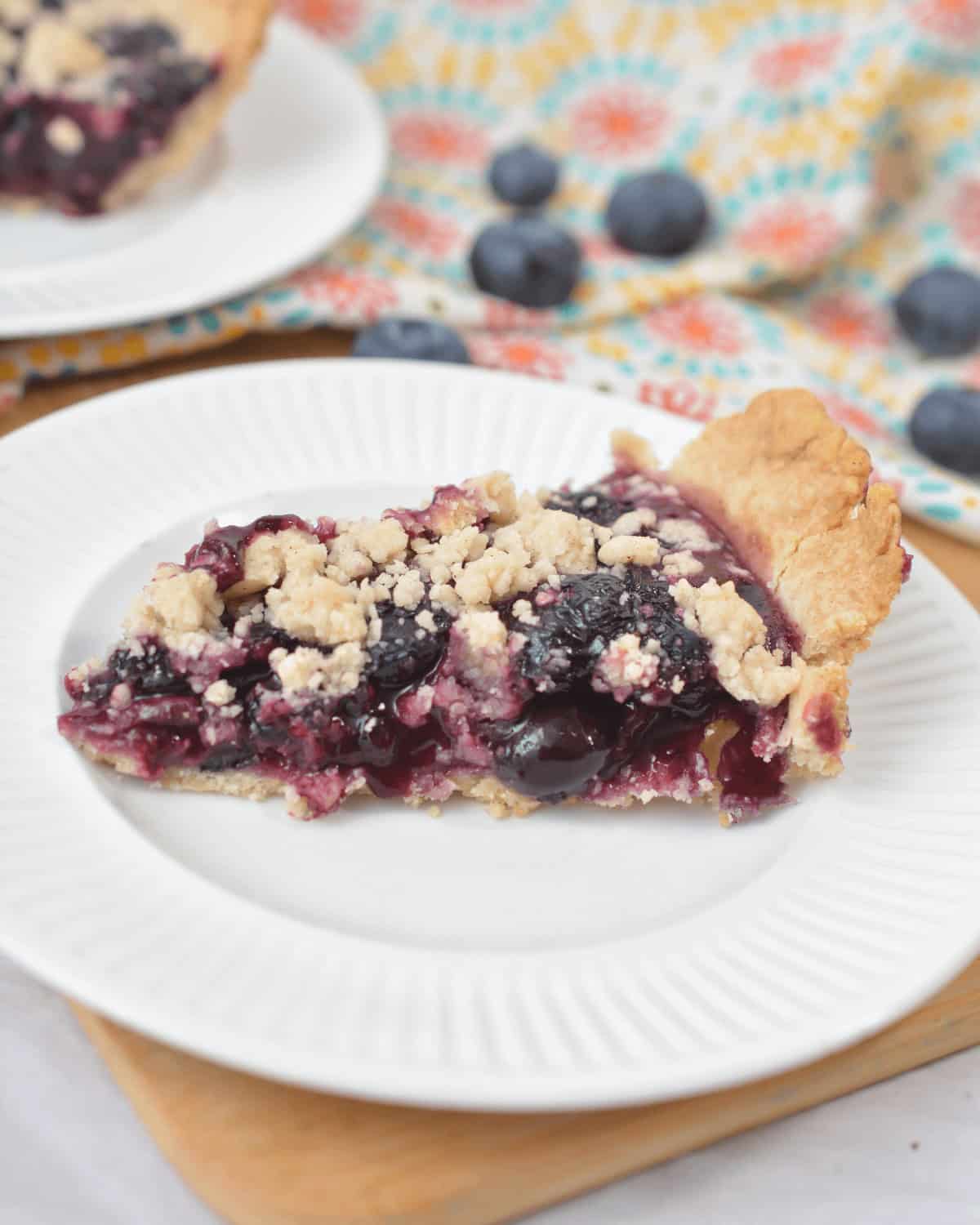 A close-up of a slice of Blueberry Crumb Pie on a white plate. The background includes a patterned cloth and scattered fresh blueberries.