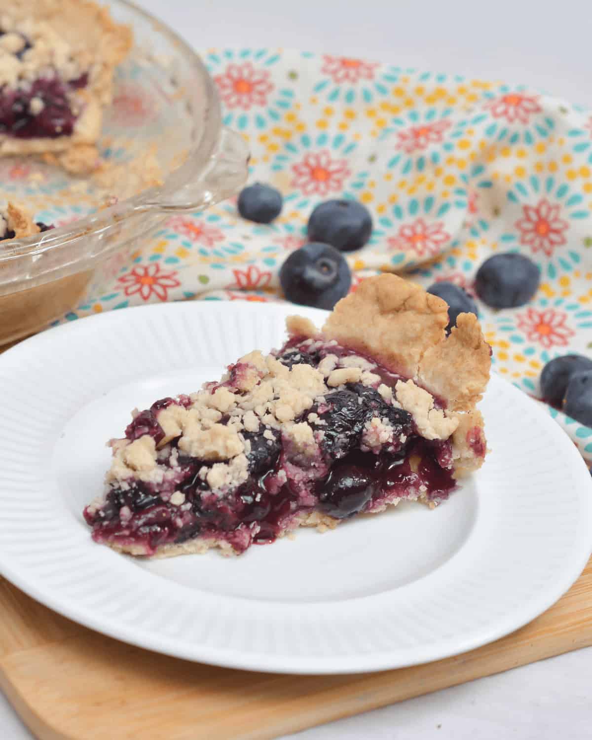 A slice of Blueberry Crumb Pie with a crumb topping sits on a white plate. A glass pie dish with remaining pie and scattered blueberries are visible in the background on a patterned cloth.