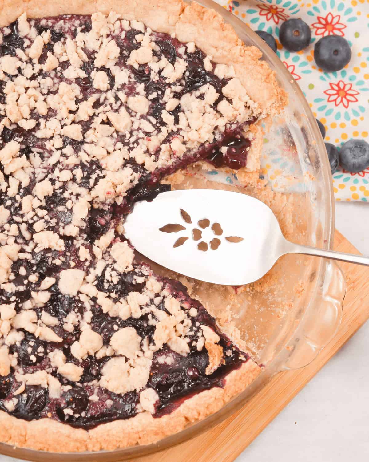 A partially eaten Blueberry Crumb Pie with a crumb topping is in a glass pie dish, placed on a wooden board. A metal pie server with a floral cutout pattern rests on the pie. Blueberries and a floral-patterned cloth are in the background.