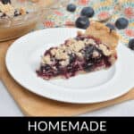 A slice of homemade Blueberry Crumb Pie on a white plate, with the remaining pie in a glass dish and a few loose blueberries around. Text reads "HOMEMADE BLUEBERRY CRUMB PIE" against a black background.
