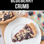 A slice of blueberry crumb pie on a white plate, with the remaining blueberry crumb pie in a baking dish nearby. Blueberries and a colorful patterned cloth are in the background.