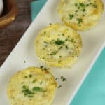 Three ham and egg cups garnished with chopped herbs are arranged in a row on a rectangular white plate, placed on a light blue napkin.