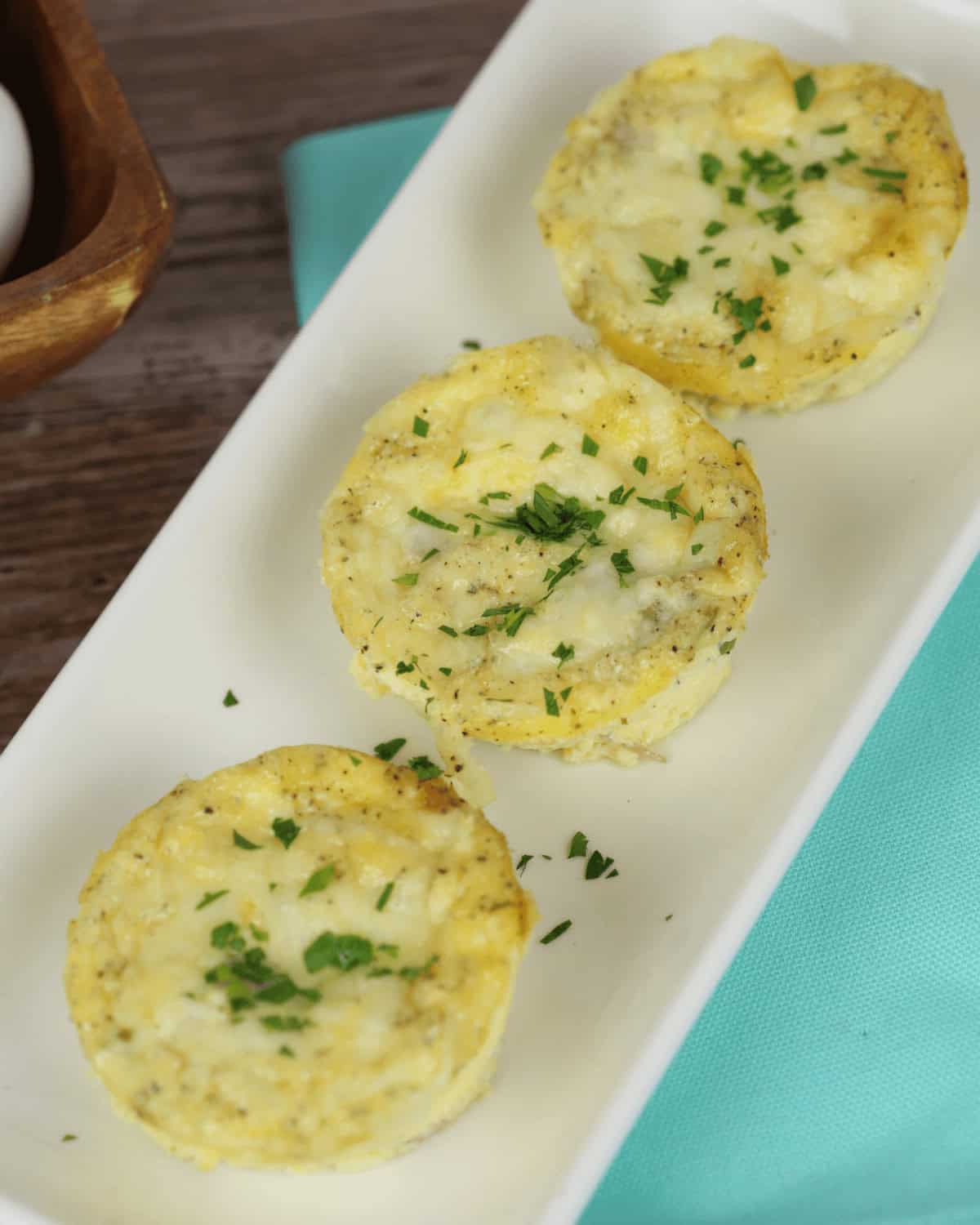 Three ham and egg cups garnished with chopped herbs are arranged in a row on a rectangular white plate, placed on a light blue napkin.