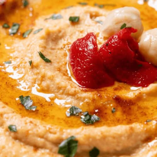 A close-up of a bowl of roasted red pepper hummus topped with chickpeas, olive oil, and parsley.