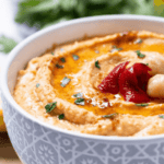 A bowl of roasted red pepper hummus garnished with herbs sits enticingly, while another bowl in the background holds vibrant red tomatoes.