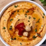 A bowl of roasted red pepper hummus garnished with chickpeas, olive oil, and parsley sits invitingly on a wooden board.