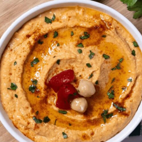 A bowl of roasted red pepper hummus garnished with chickpeas, olive oil, and parsley sits invitingly on a wooden board.