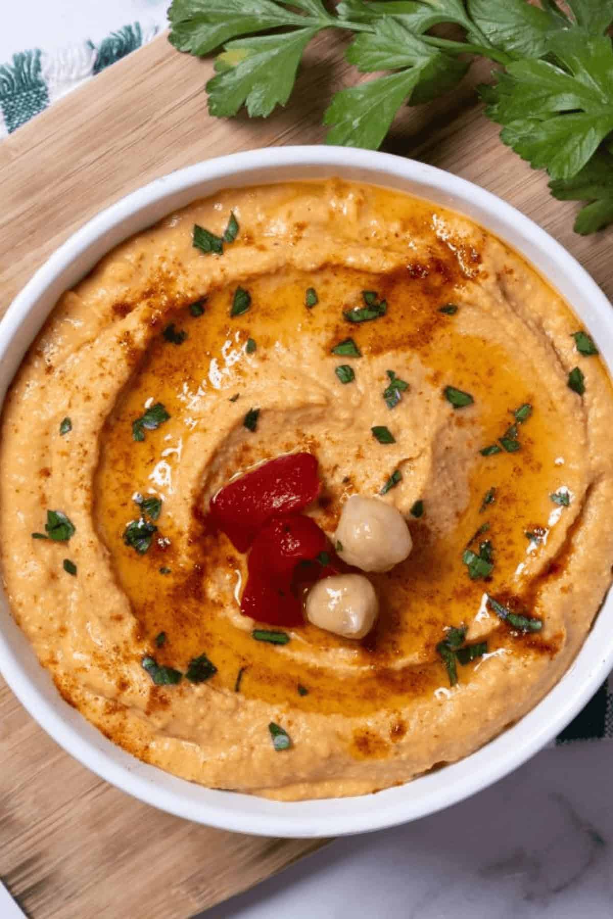 A bowl of roasted red pepper hummus garnished with chickpeas, olive oil, and parsley sits invitingly on a wooden board.