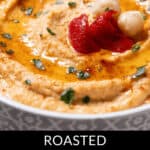 A close-up of a bowl of roasted red pepper hummus garnished with chickpeas, herbs, and vibrant red pepper pieces creates an inviting display.