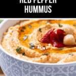 A bowl of roasted red pepper hummus, elegantly garnished with fresh herbs, vibrant red peppers, and plump chickpeas on top.