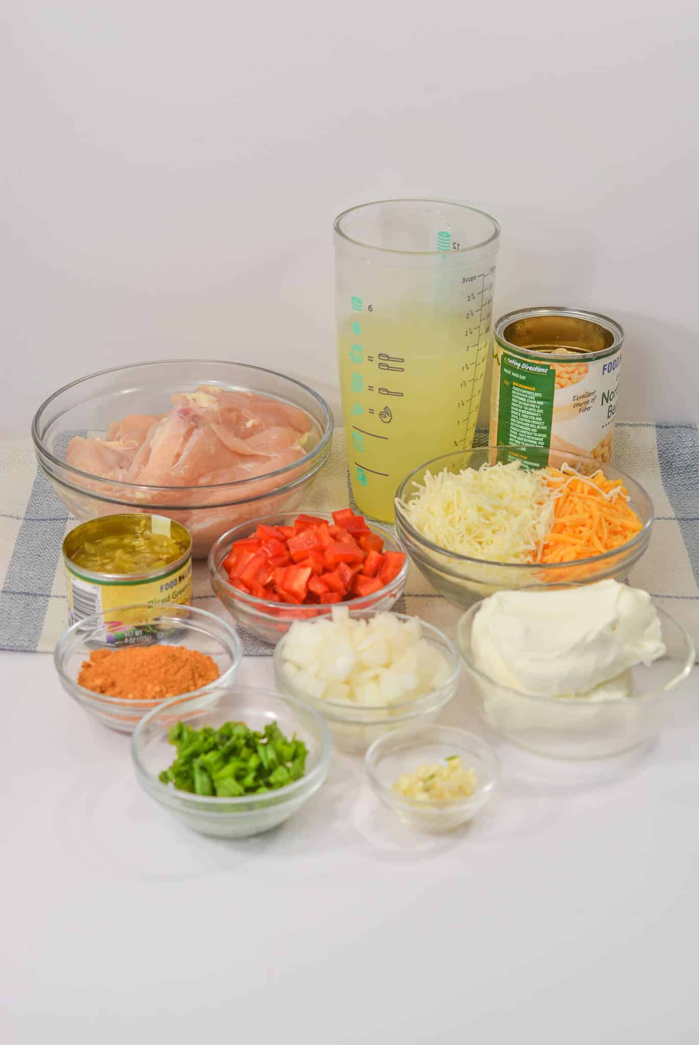 Various ingredients are arranged on a table, ready for Cream Cheese Chicken Chili. You'll find raw chicken, canned goods, cheese, diced tomatoes, onions, green onions, cream cheese, and a measuring cup with liquid—all set to create a cozy meal.
