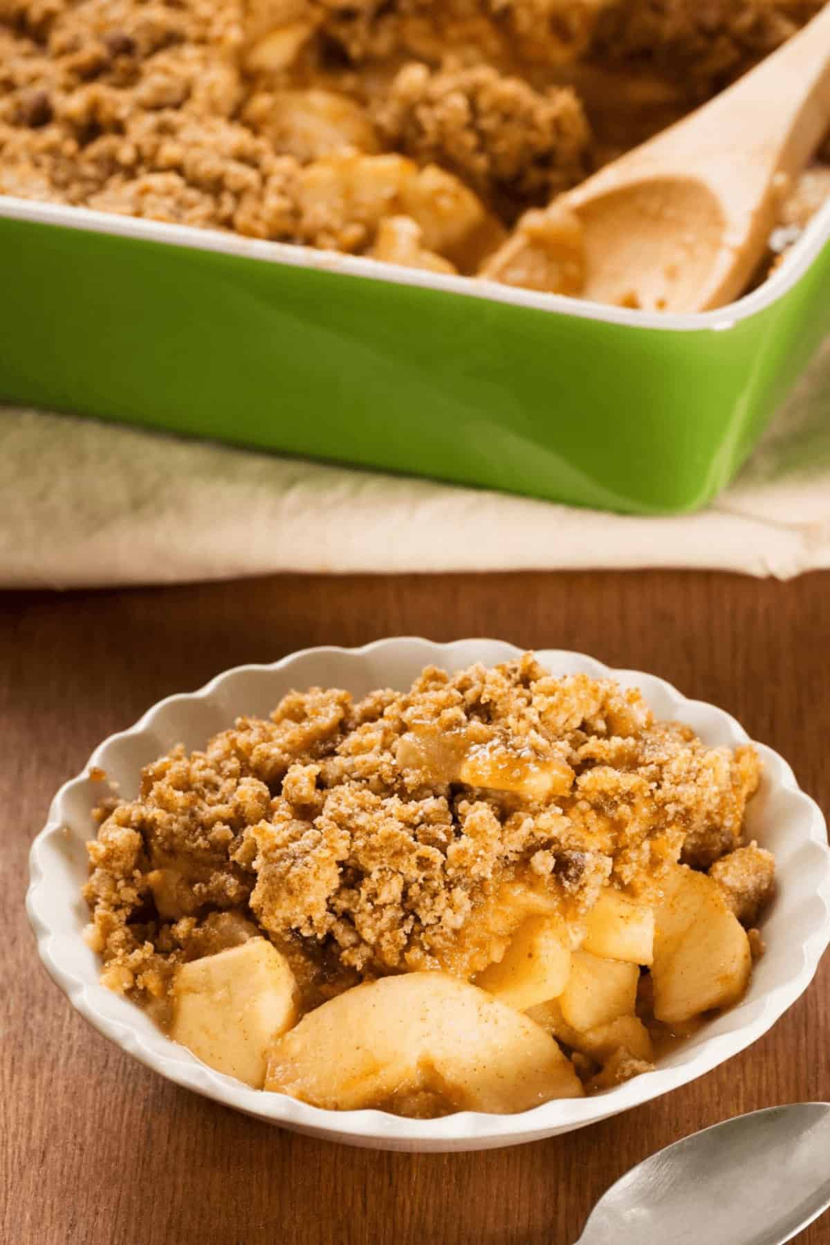 A bowl of easy apple crumble with a crumbly topping is in the foreground. A baking dish with more apple crisp and a wooden spoon is in the background, promising effortless indulgence.