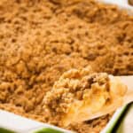 A wooden spoon lifts a portion of easy apple crumble from a green baking dish. The dessert has a crumbly topping and appears golden brown, making it an effortless delight for any occasion.