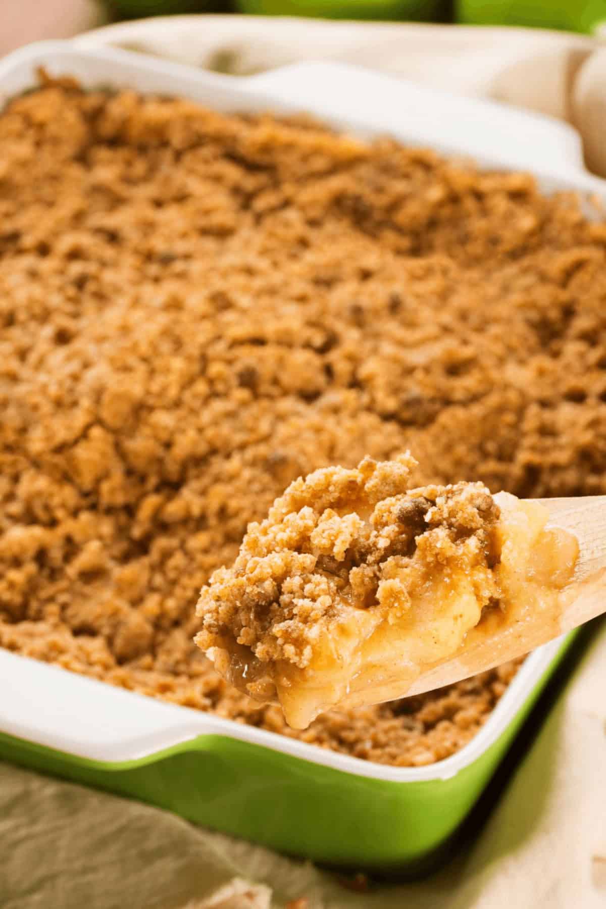 A wooden spoon lifts a portion of easy apple crumble from a green baking dish. The dessert has a crumbly topping and appears golden brown, making it an effortless delight for any occasion.