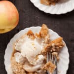 Two white plates present servings of easy apple crumble, each topped with a scoop of vanilla ice cream, next to a bright whole apple on a dark wooden surface.