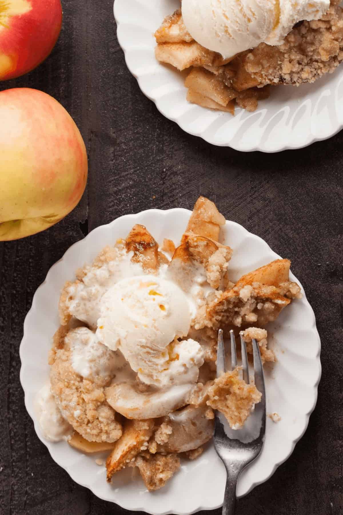 Two white plates present servings of easy apple crumble, each topped with a scoop of vanilla ice cream, next to a bright whole apple on a dark wooden surface.