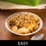 A white dish with a serving of Easy Apple Crumble sits invitingly in front of a green baking dish. A spoon rests beside it, ready to delve into this delightful dessert. Text reads: "Easy Apple Crumble.