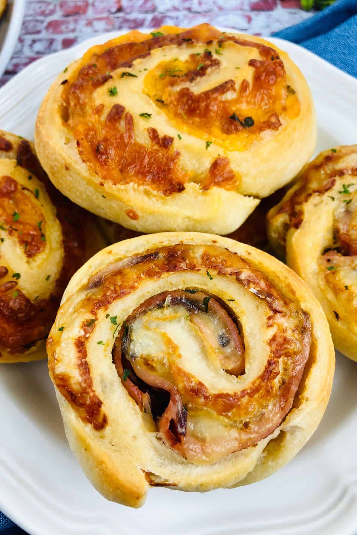 A close-up of a white plate holding several baked Ham and Cheese Roll Ups, their golden pinwheel layers revealing melted cheese and herbs.