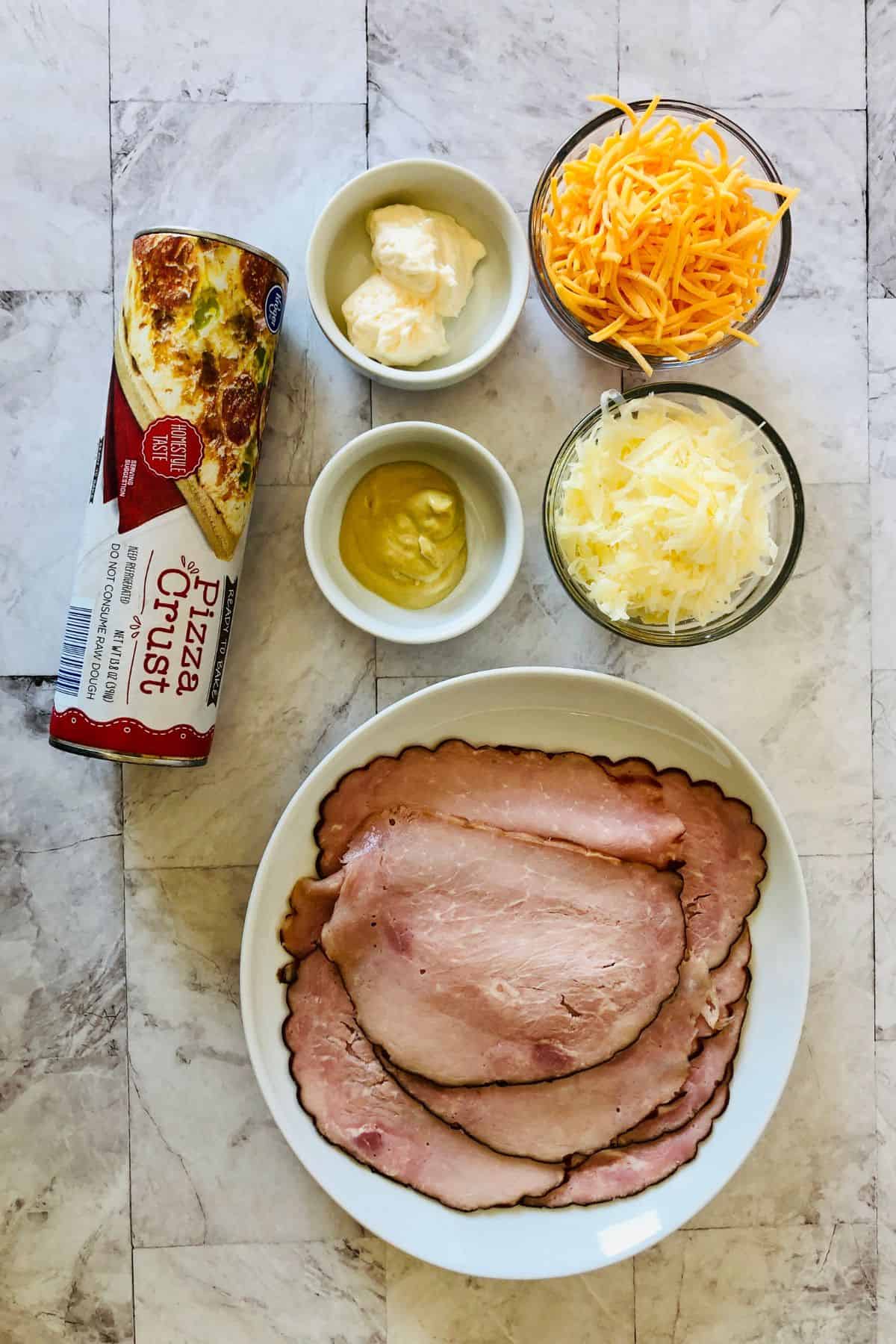 A plate with sliced ham, a can of pizza crust, bowls of shredded cheddar cheese, shredded mozzarella, mayonnaise, and mustard are arranged on a light surface—perfect ingredients for making delicious Ham and Cheese Roll Ups.