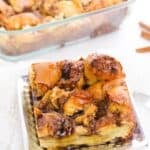 A square piece of cinnamon roll casserole on a plate, with the rest of the Brioche French Toast Casserole in a glass baking dish in the background.