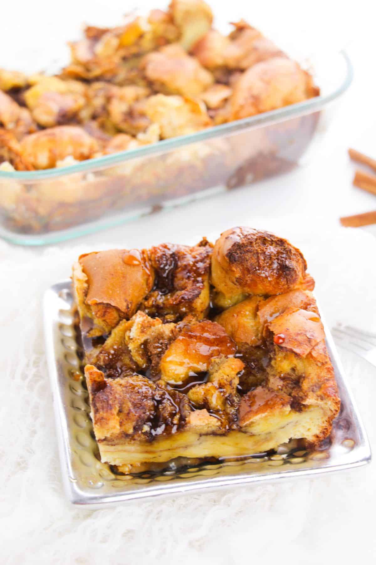 A square piece of cinnamon roll casserole on a plate, with the rest of the Brioche French Toast Casserole in a glass baking dish in the background.