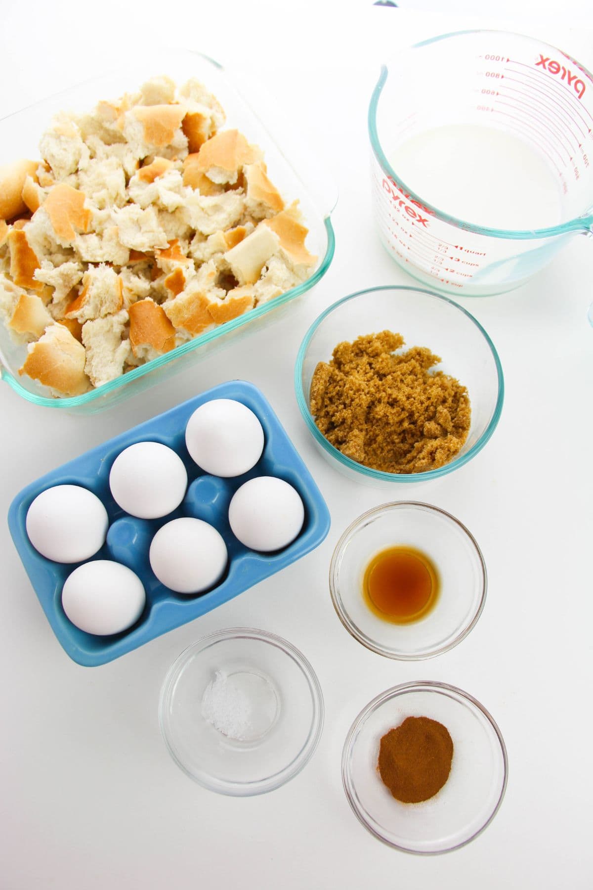 Glass containers with torn bread, milk, brown sugar, vanilla extract, eggs, salt, and cinnamon are arranged on a white surface.