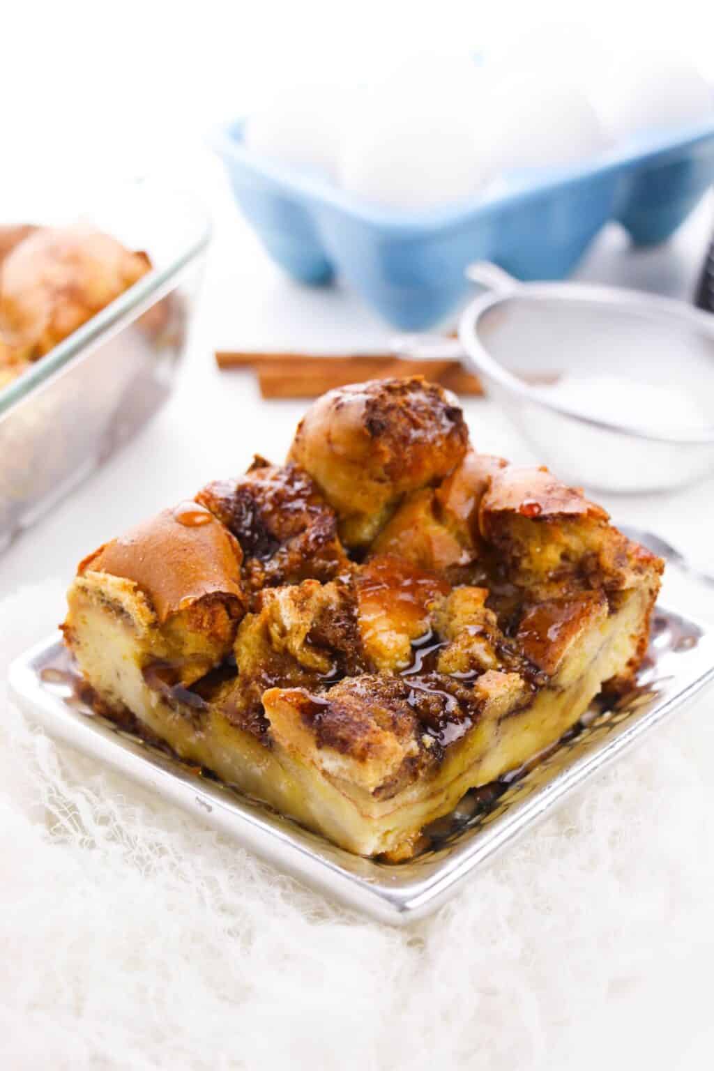 A square serving of bread pudding with a caramelized top sits on a silver plate, reminiscent of Brioche French Toast Casserole; eggs, cinnamon sticks, and a baking dish are in the background.