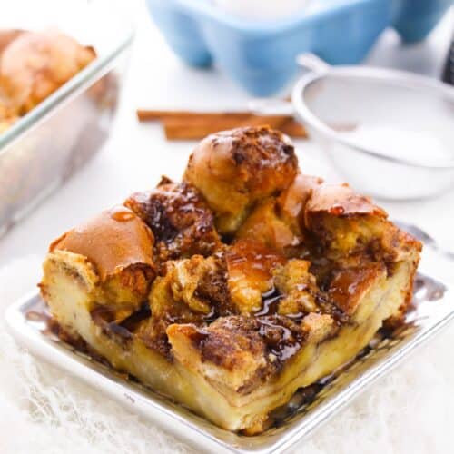 A square serving of bread pudding with a caramelized top sits on a silver plate, reminiscent of Brioche French Toast Casserole; eggs, cinnamon sticks, and a baking dish are in the background.