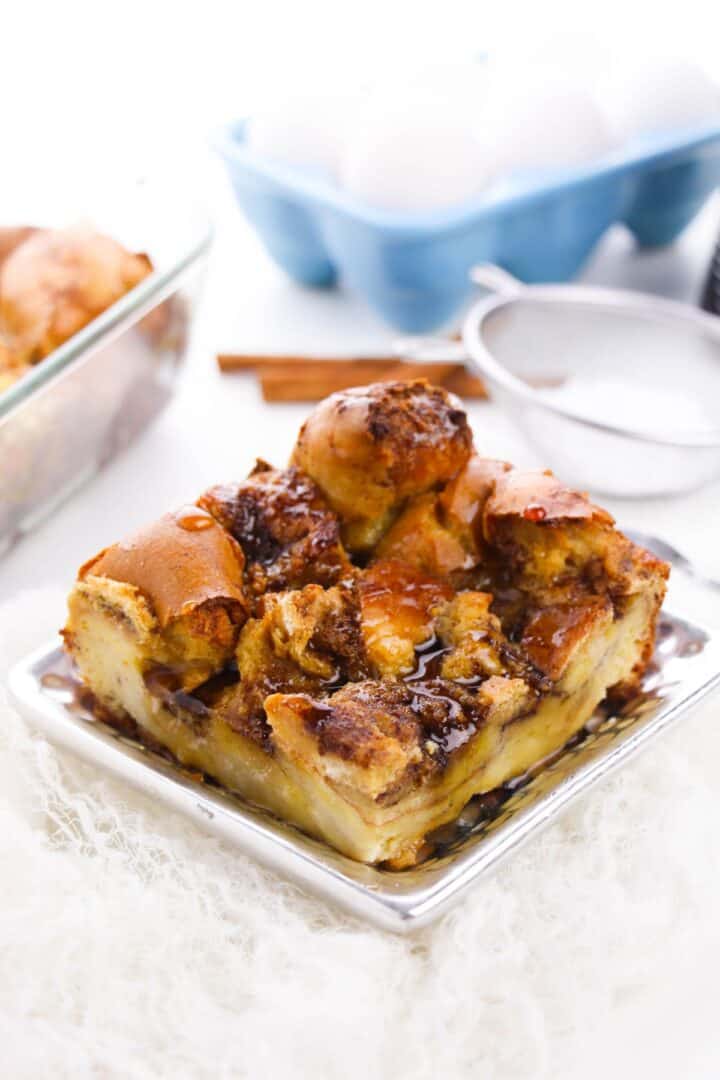 A square serving of bread pudding with a caramelized top sits on a silver plate, reminiscent of Brioche French Toast Casserole; eggs, cinnamon sticks, and a baking dish are in the background.