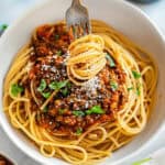 A bowl of pasta topped with bolognese, fresh basil, and parmesan cheese, with a fork twirling the pasta.